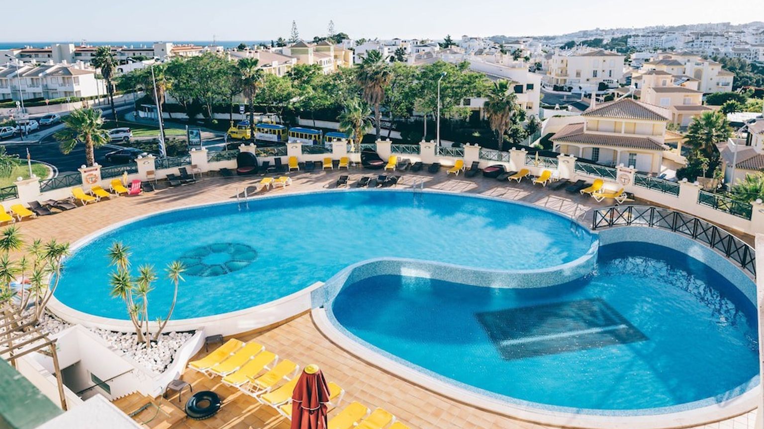 En flygbild av ett resortkomplex med två blå, sammanlänkade pooler omgivna av många gula solstolar och bostadsbyggnader i bakgrunden.