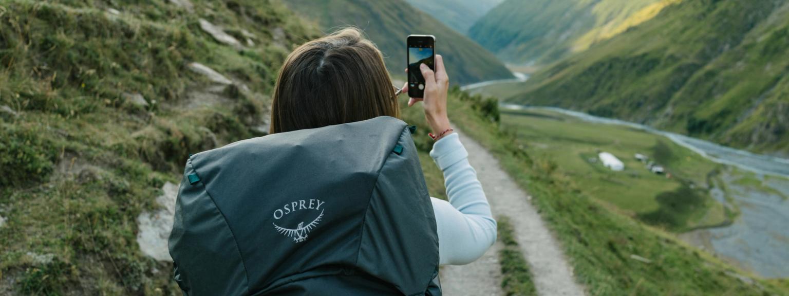 en kvinna med en osprey-ryggsäck tar en bild av ett berg med sin mobiltelefon