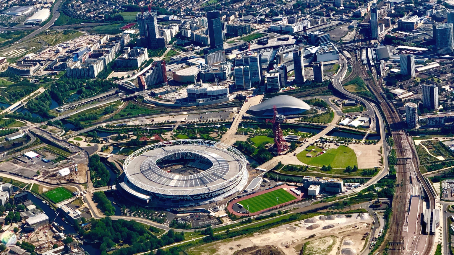 Aerial view of the London Olympic Park, featuring the main stadium, other venues, green spaces, and surrounding city.