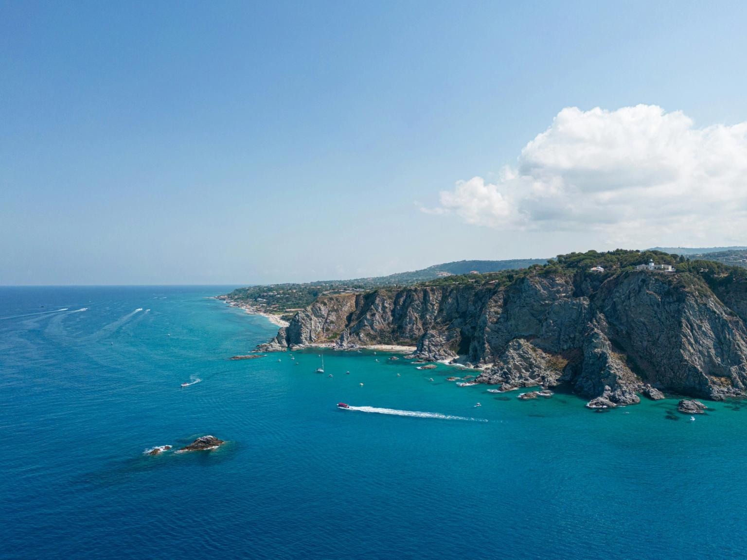Veduta aerea di una costa vibrante con scogliere frastagliate, una spiaggia sabbiosa e acqua azzurra e limpida con barche. Capo Vaticano, Italia