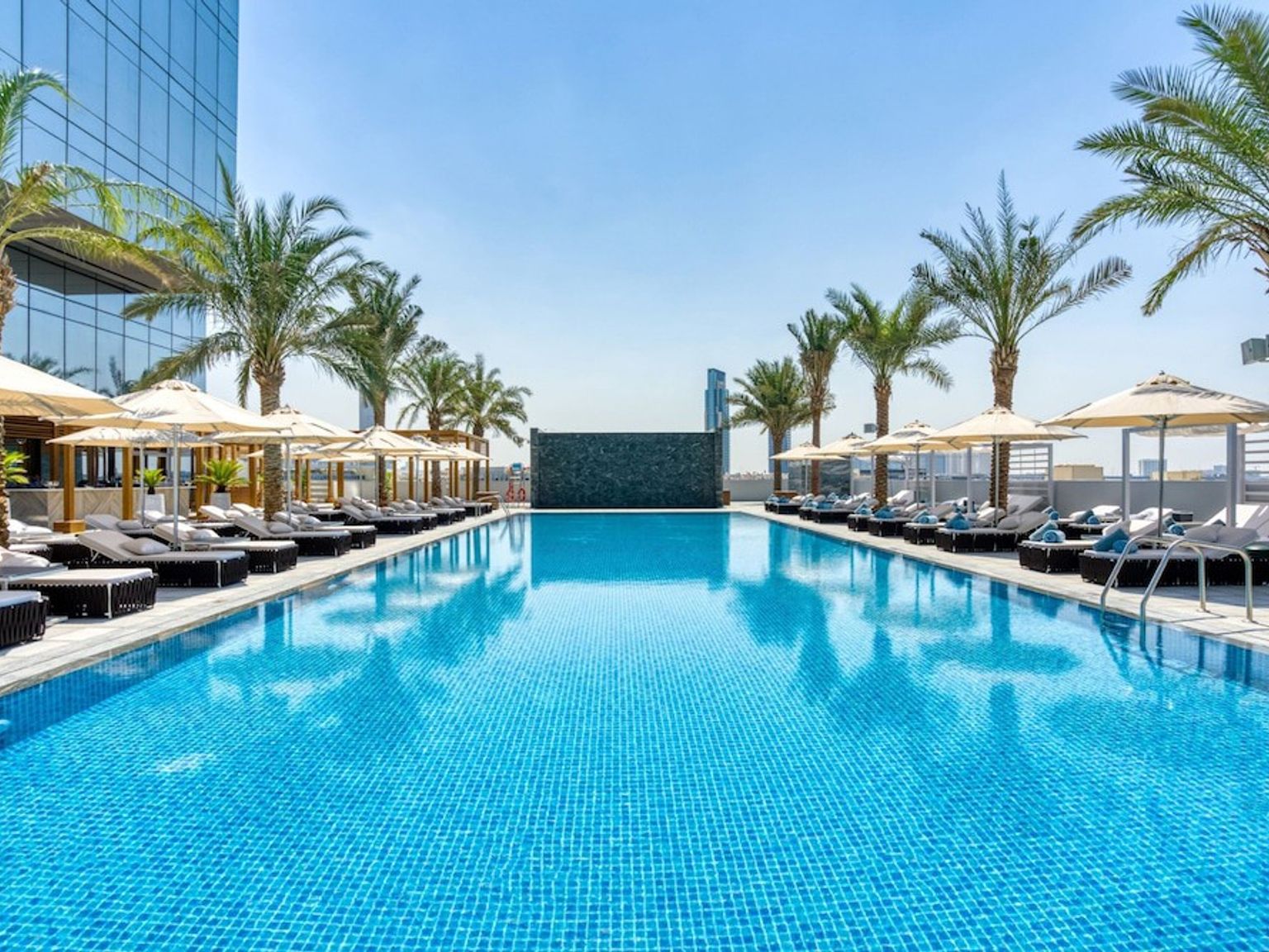 Lang utendørs svømmebasseng flankert av solsenger, parasoller og palmer.