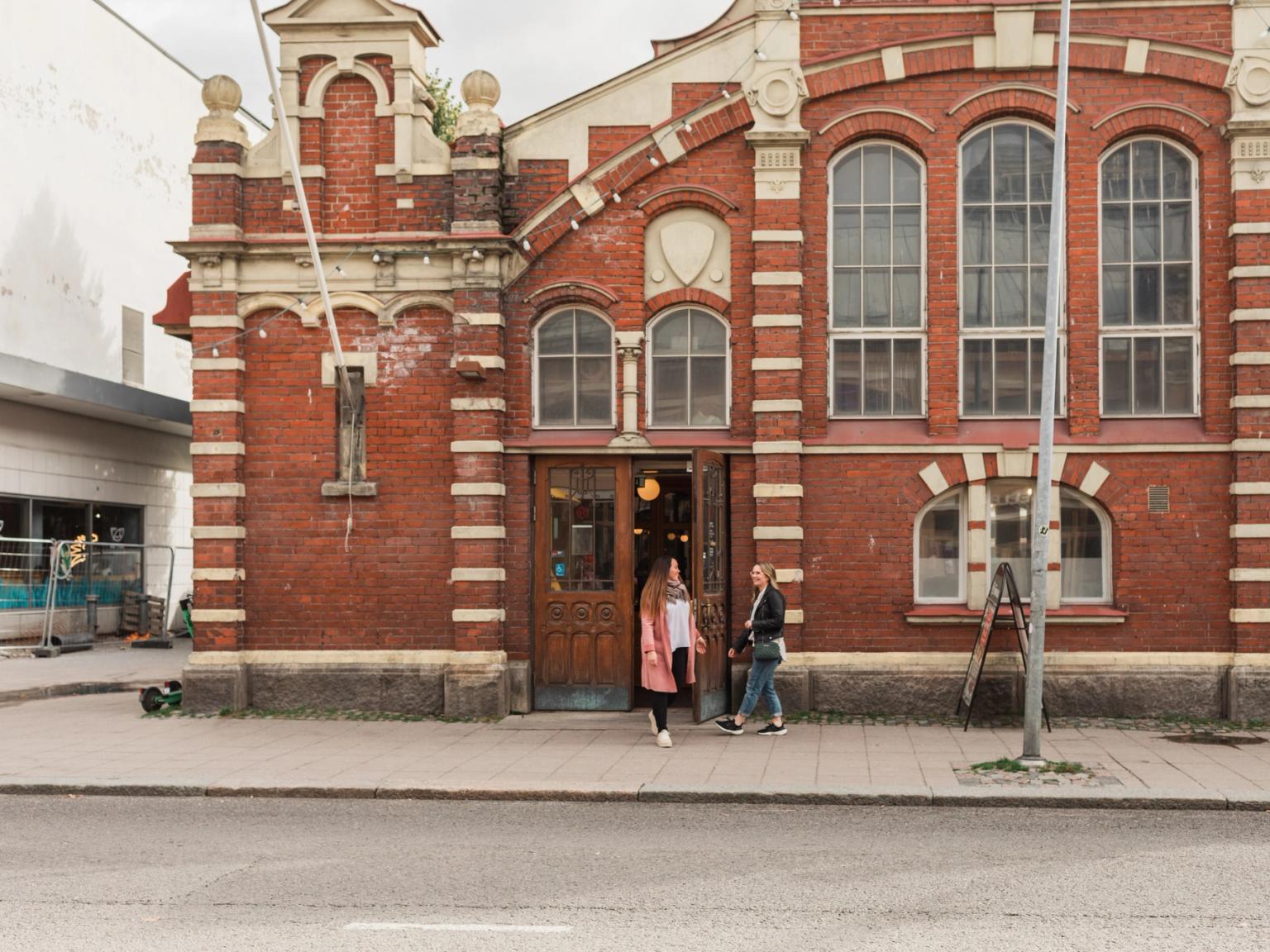 Koristeellinen punatiilirakennus, jossa on valkoiset reunat, kaksi naista seisoo sen sisäänkäynnillä kaupunkikadulla.