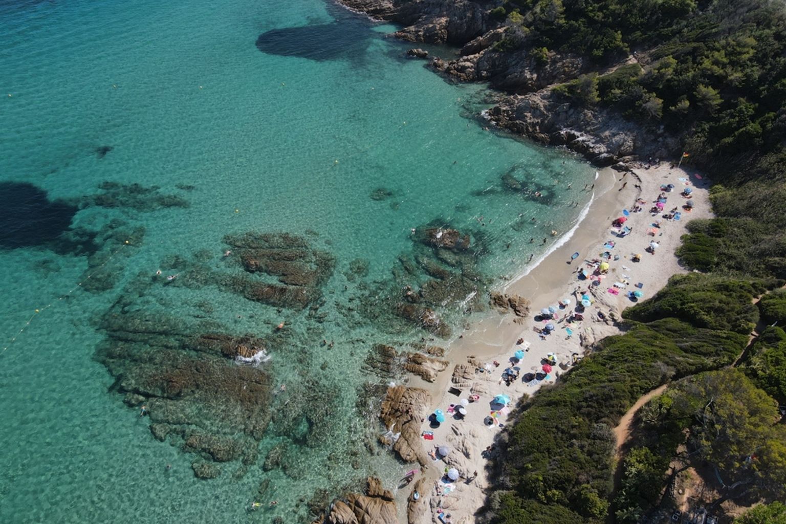Aerial view of a crowded sandy beach with colorful umbrellas nestled in a rocky cove with clear turquoise water. Cap Taillat, France