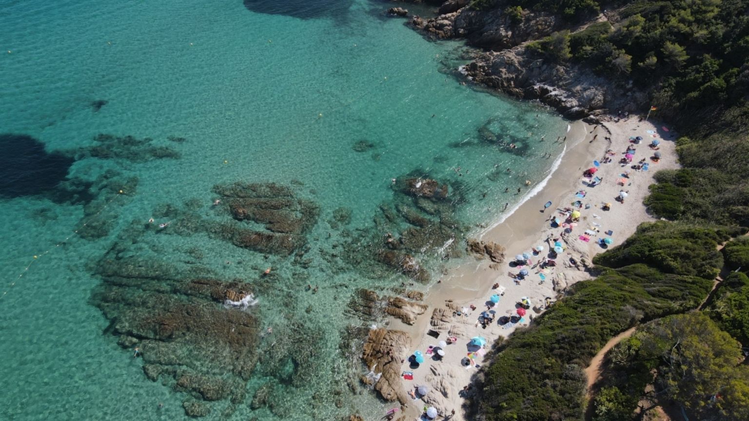 Aerial view of a crowded sandy beach with colorful umbrellas nestled in a rocky cove with clear turquoise water. Cap Taillat, France
