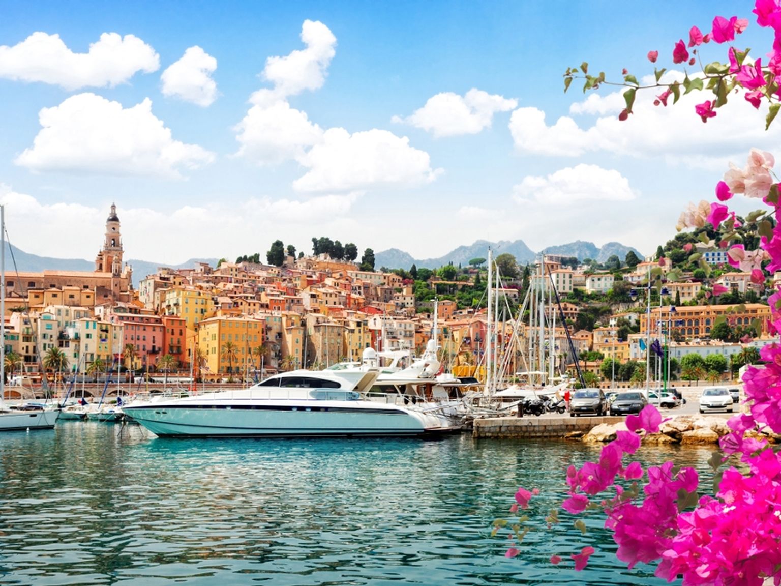Kuvankaunis satamakohtaus, jossa on värikkäitä rakennuksia, jahteja, kirkontorni ja elinvoimainen pinkki bougainvillea. Menton, Ranska