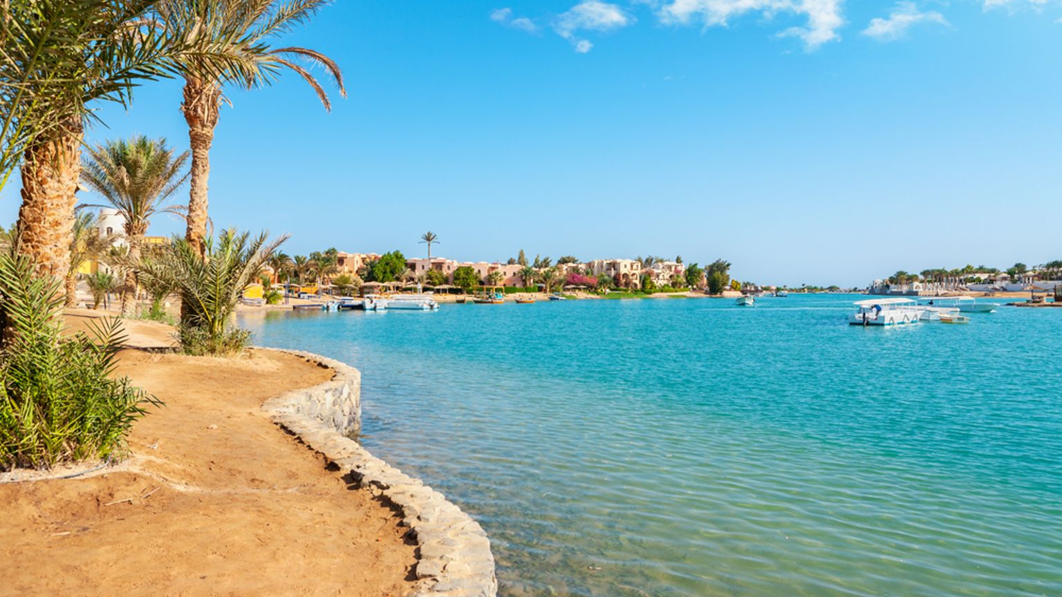 Palmer och en sandbank kantar en turkos kanal med båtar och resortbyggnader under en blå himmel. El Gouna, Egypten
