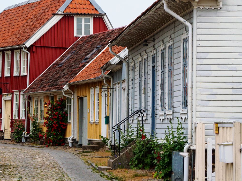 En række farverige træhuse i rød, gul og lyseblå langs en brostensbelagt gade med grønne omgivelser.