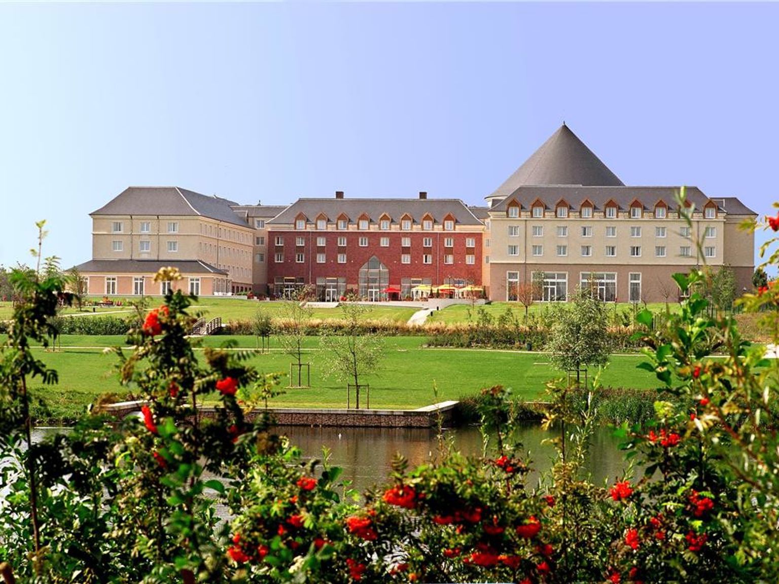 Et stort hotell med krem- og røde seksjoner, et konisk tak, en grønn plen og en dam med busker med røde bær. Grand Magic Hotel