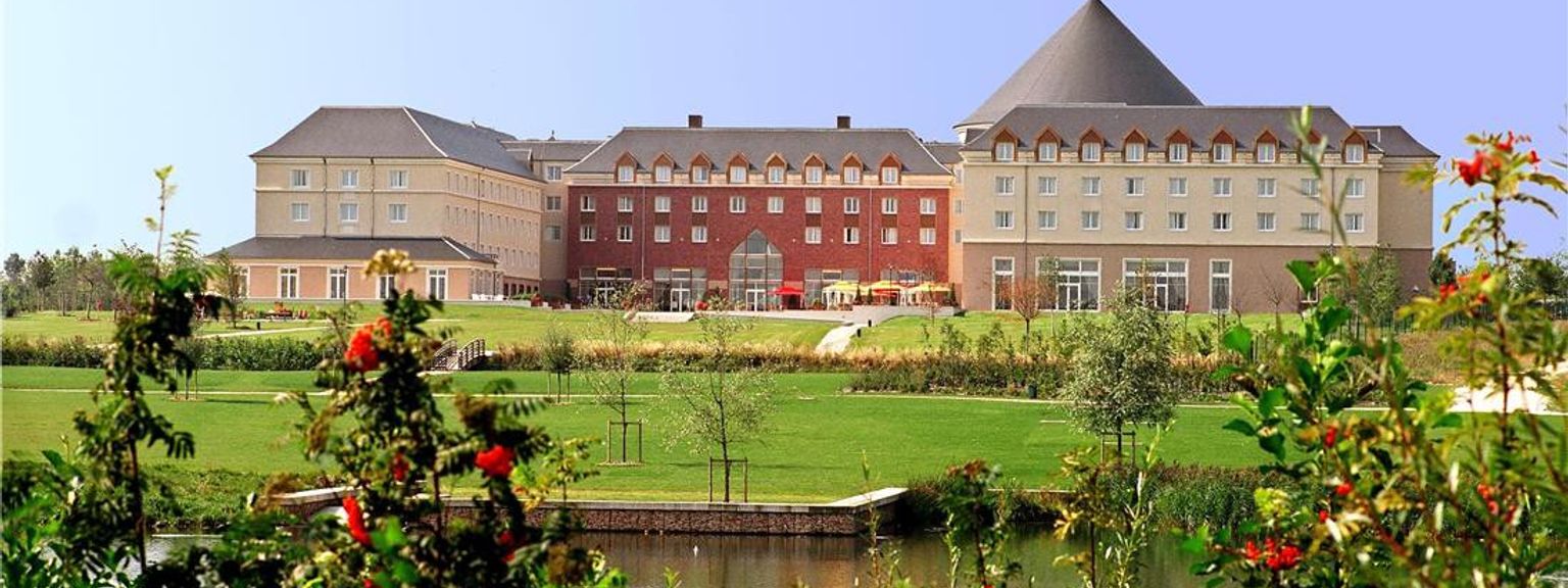 Et stort hotell med krem- og røde seksjoner, et konisk tak, en grønn plen og en dam med busker med røde bær. Grand Magic Hotel