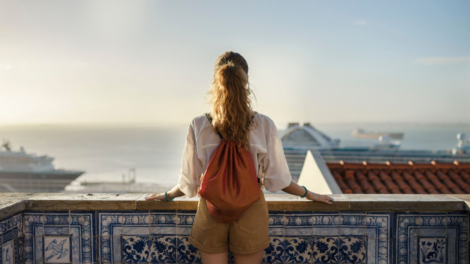 En kvinna med ryggsäck, bakifrån, lutar sig mot ett blått kakelräcke med utsikt över en stadshamn med fartyg.