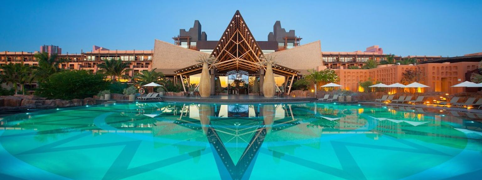 Stor resortpool, der reflekterer en karakteristisk A-ramme indgangsstruktur ved skumringen, med månen på himlen.