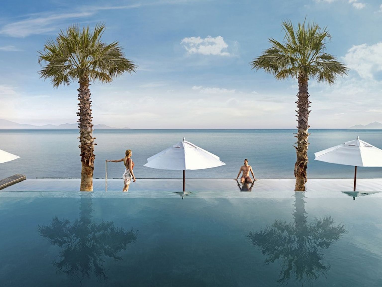 två personer sitter i en simbassäng bredvid palmträd med utsikt över havet.