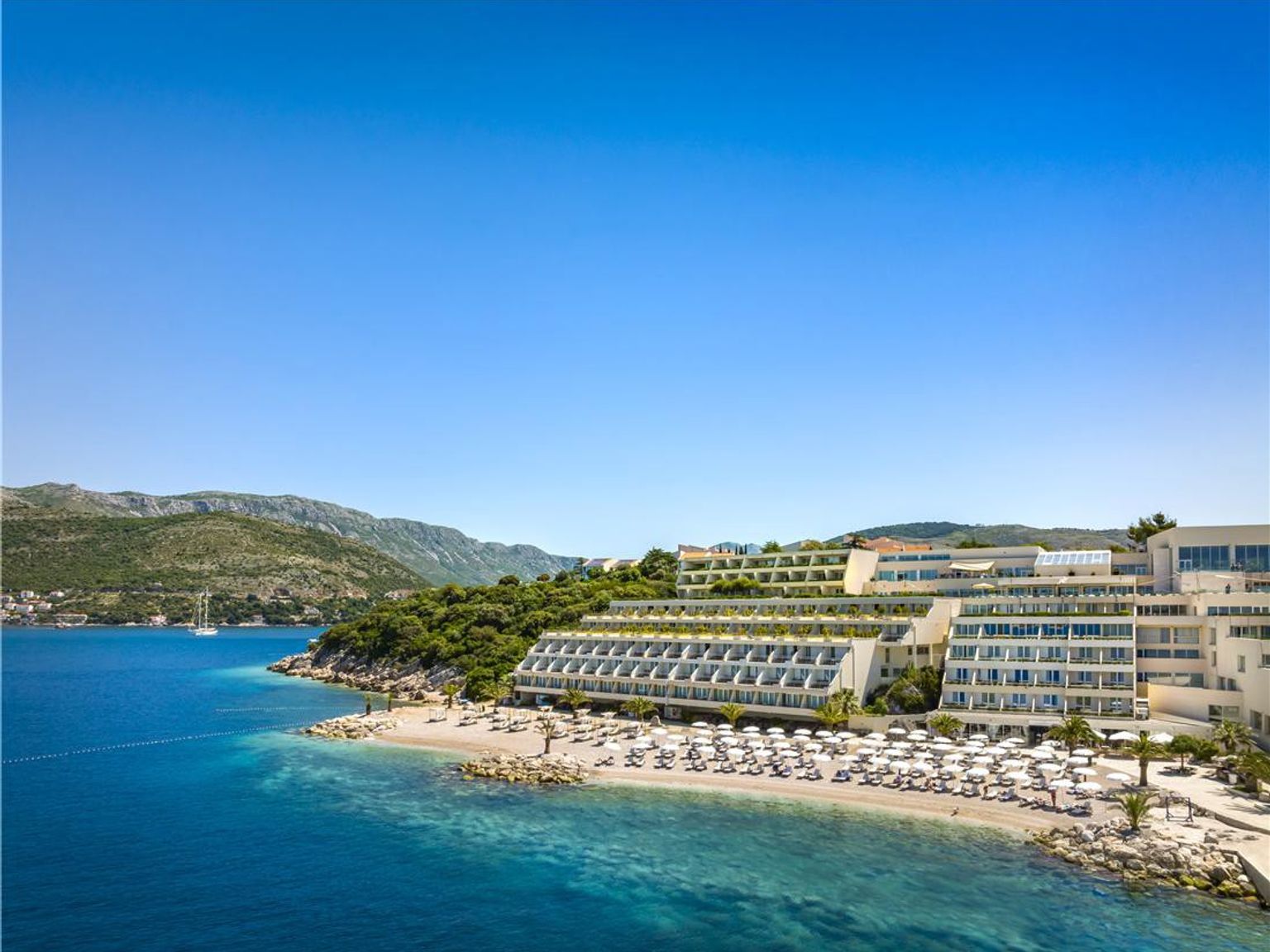 Modernt terrasshotell på en strand bredvid en blå bukt, med berg i fjärran. Dubrovnik President Valamar Collection Hotel