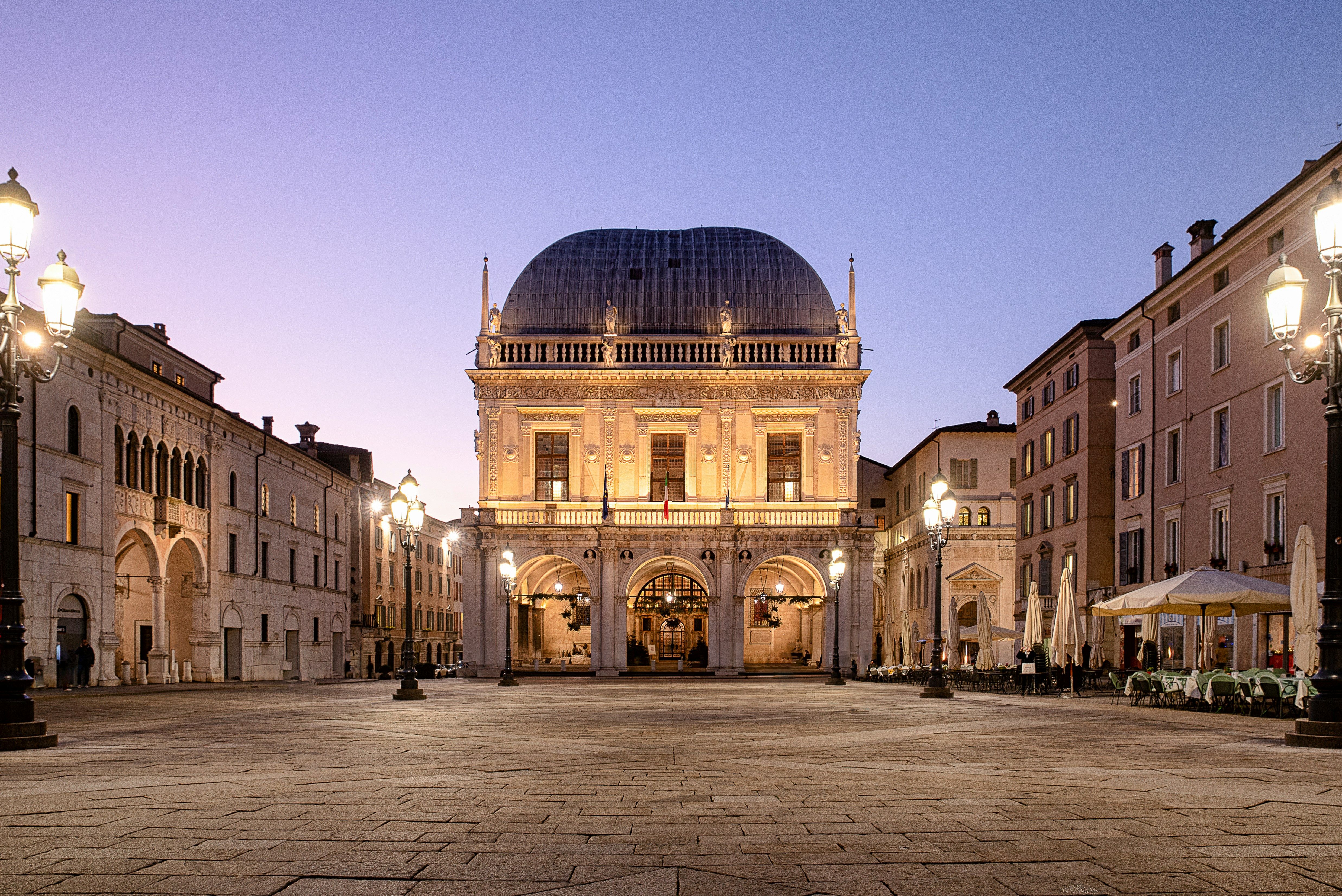 Upplyst historiskt torg i skymningen med en framträdande kupolbyggnad och många utsmyckade gatlyktor. Brescia, Italien