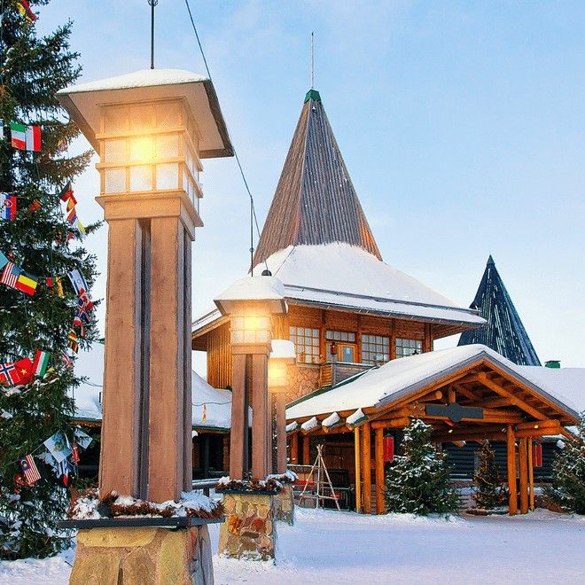 Snötäckta trähus med spetsiga tak och upplysta gatlyktor, bredvid en julgran prydd med flaggor. Jultomtens besök, Rovaniemi
