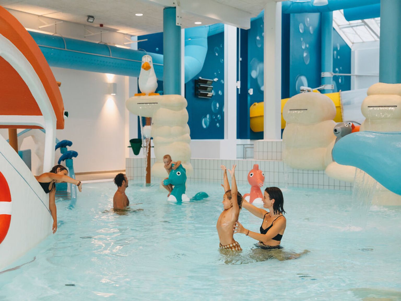 Voksne og barn nyter en fargerik innendørs vannpark med lekne strukturer og sklier.