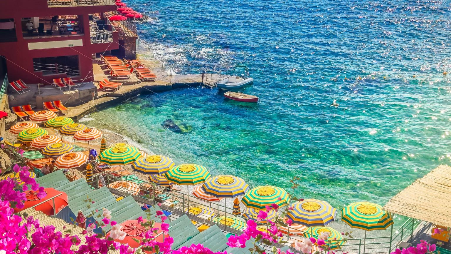 Plage animée à Capri avec des fleurs roses, des parasols rayés, une eau bleu clair et un bâtiment en terrasses.