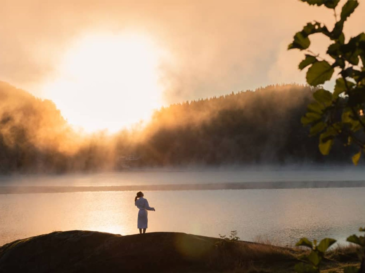 en kvinne står på en stein nær en innsjø ved solnedgang
