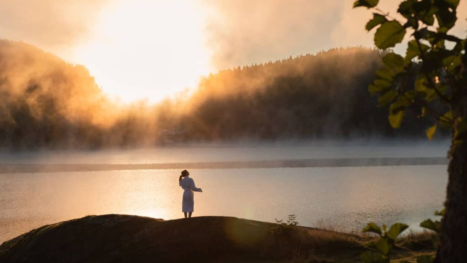 en kvinna står på en klippa nära en sjö vid solnedgången
