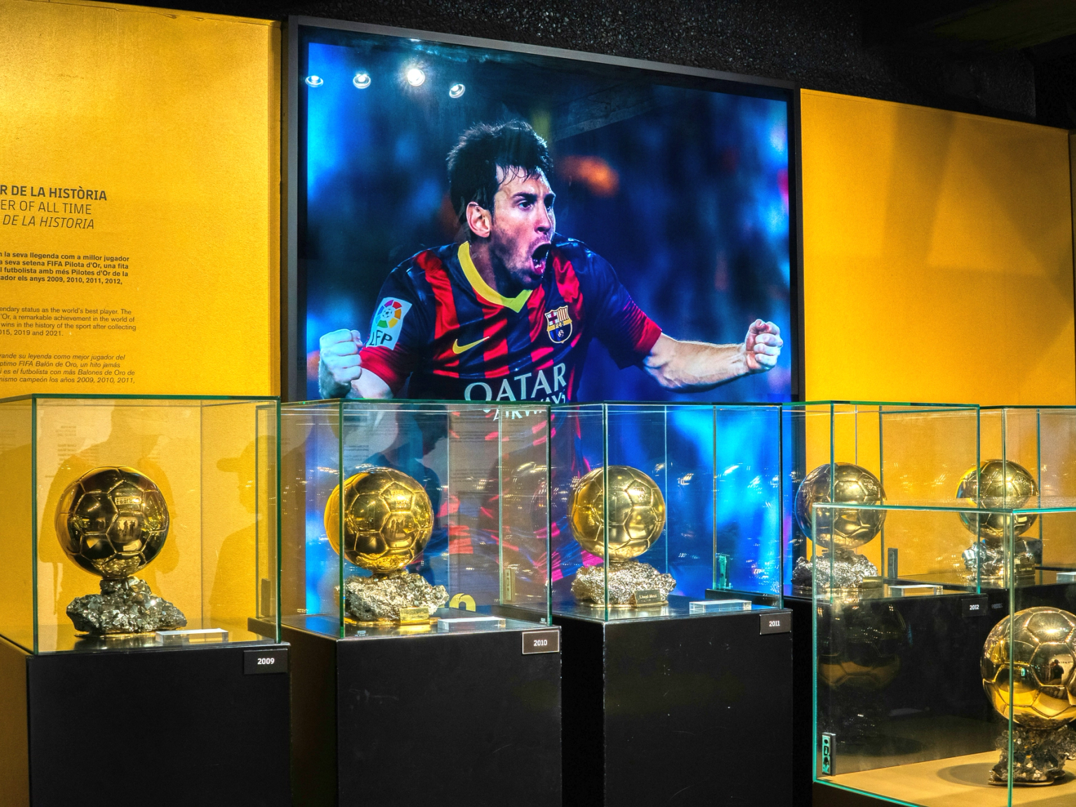 Mehrere goldene Fußballtrophäen in Glaskästen unter einem großen Bildschirm, der Lionel Messi beim Feiern zeigt.