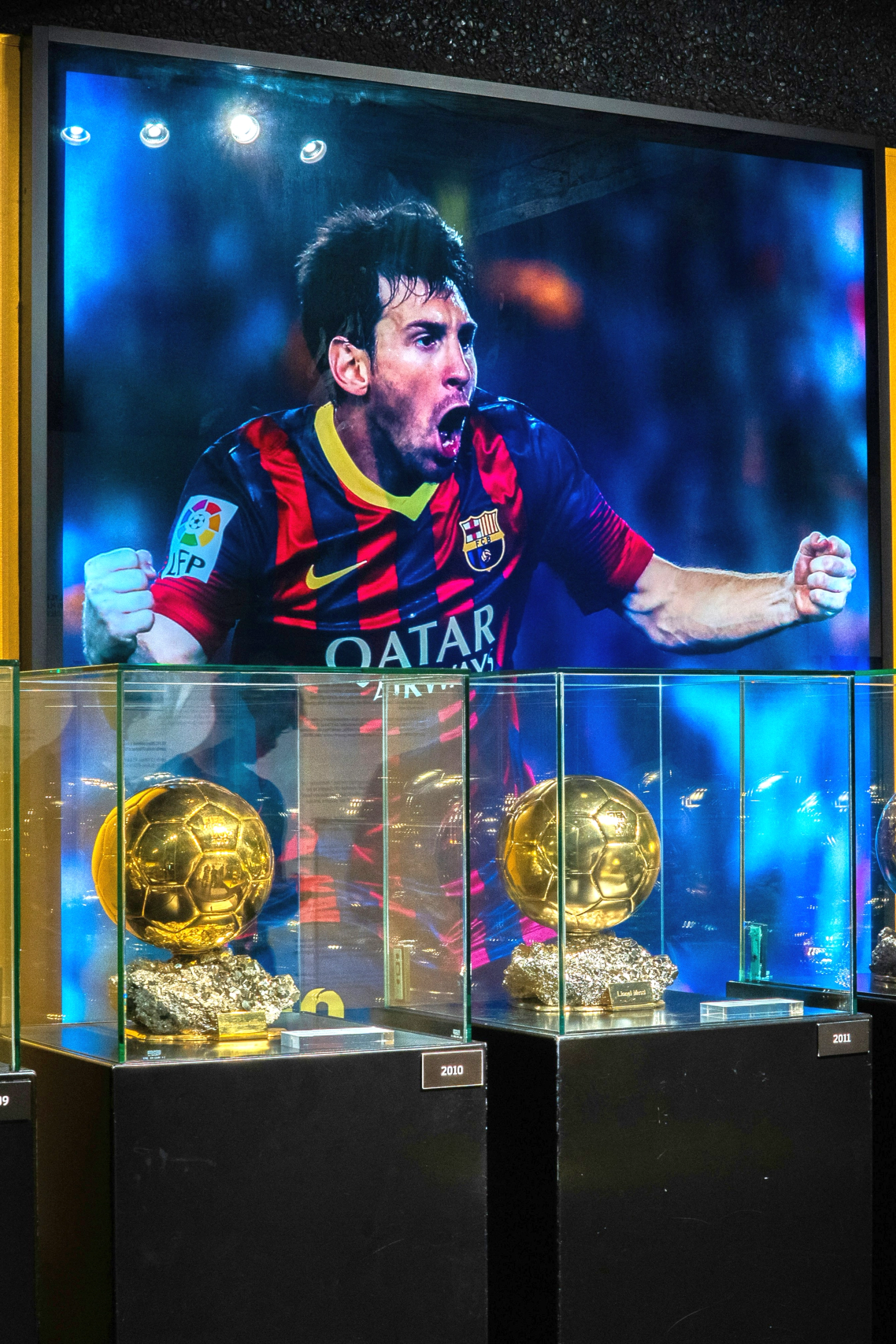 Mehrere goldene Fußballtrophäen in Glaskästen unter einem großen Bildschirm, der Lionel Messi beim Feiern zeigt.