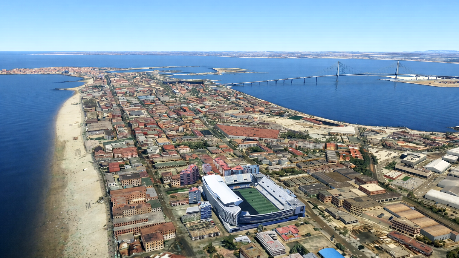 Aerial view of a coastal city on a peninsula, featuring a stadium, a beach, and a cable-stayed bridge spanning a bay.