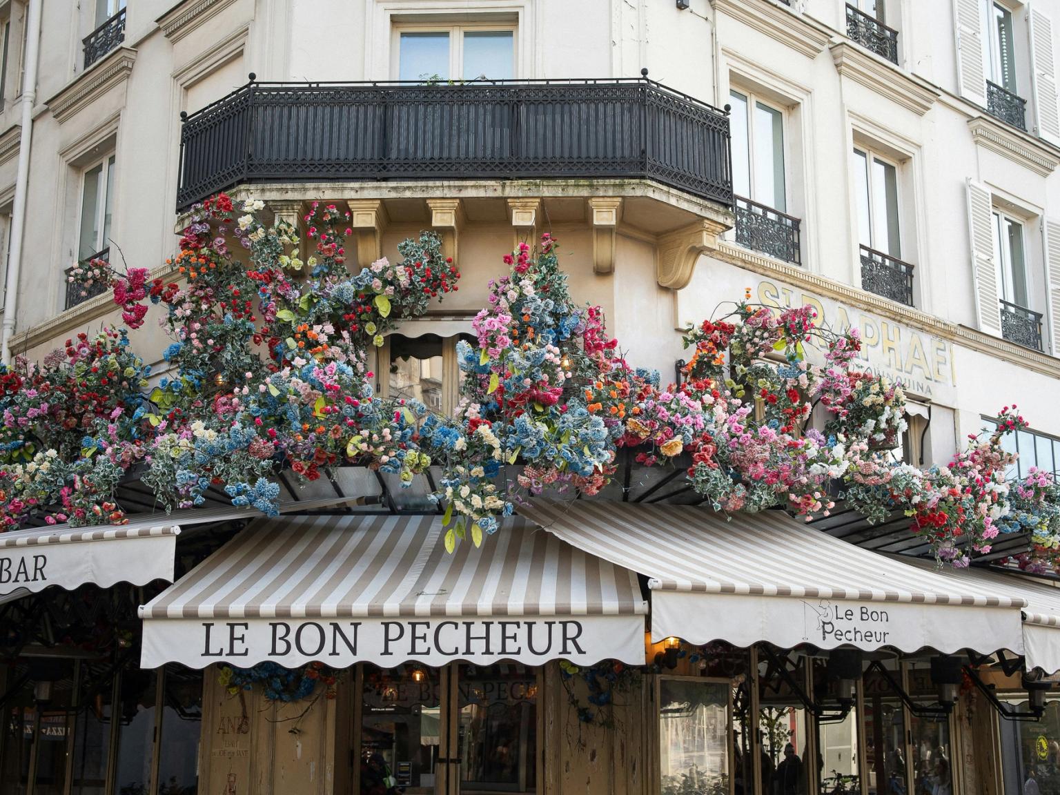 Le Bon Pecheur café on a building corner, with striped awnings overflowing with colorful artificial flowers. Paris