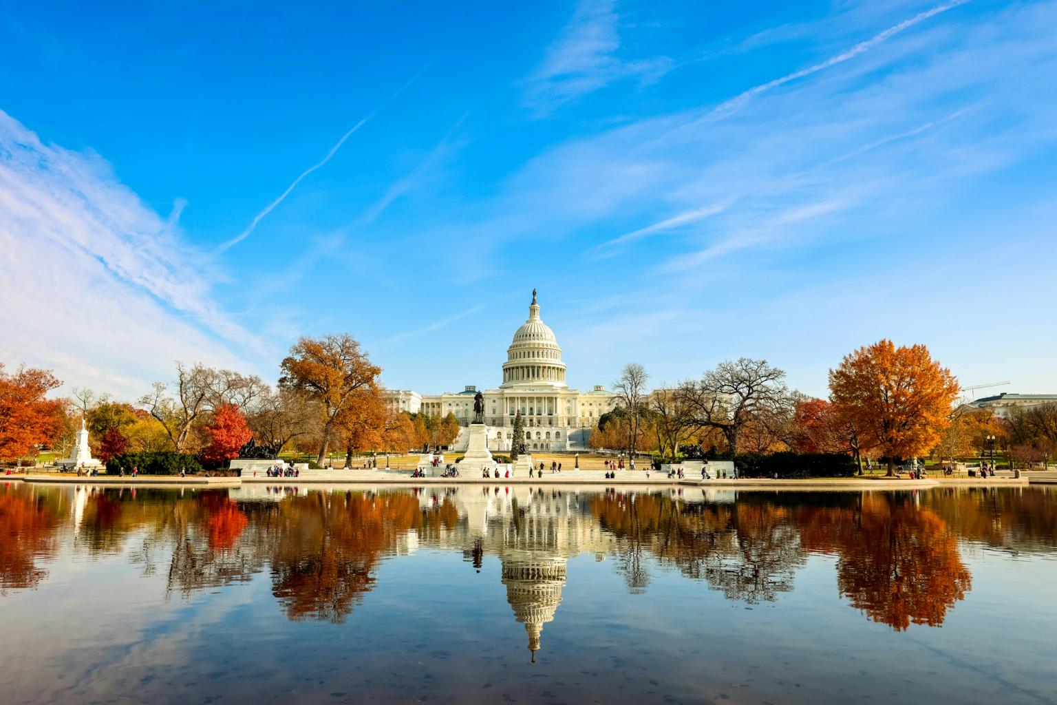 USA's Capitol-bygning spejler sig i et bassin med efterårstræer under en blå himmel.
