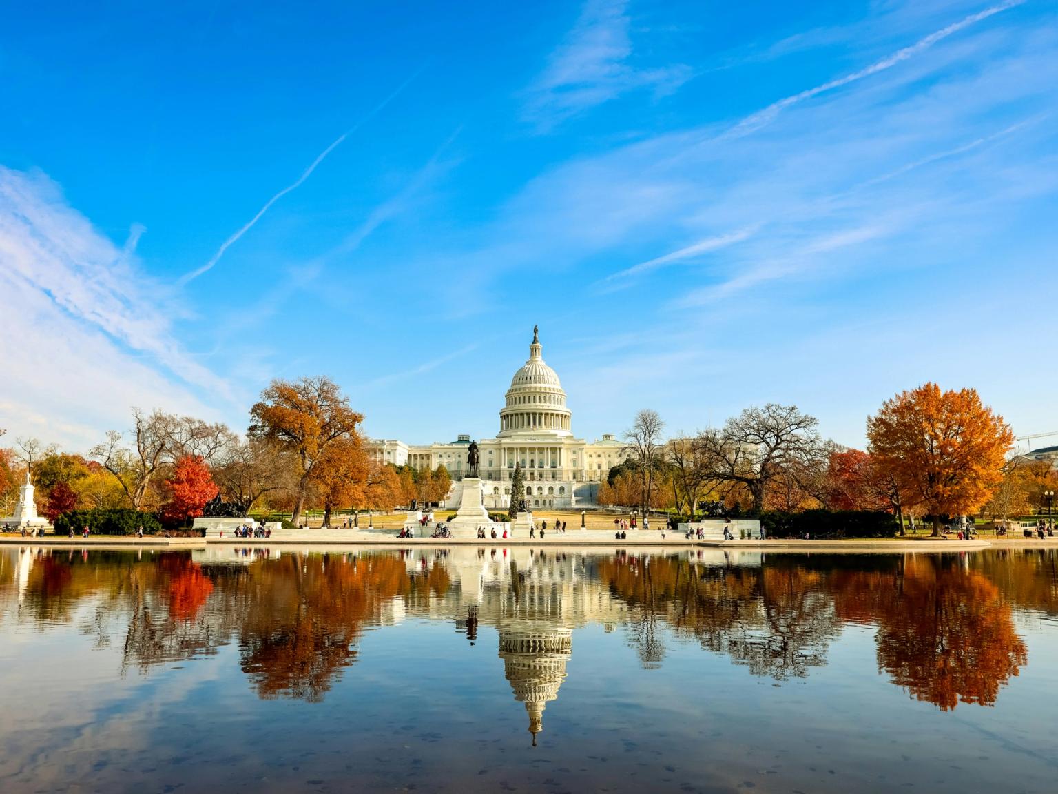 USA's Capitol-bygning spejler sig i et bassin med efterårstræer under en blå himmel.