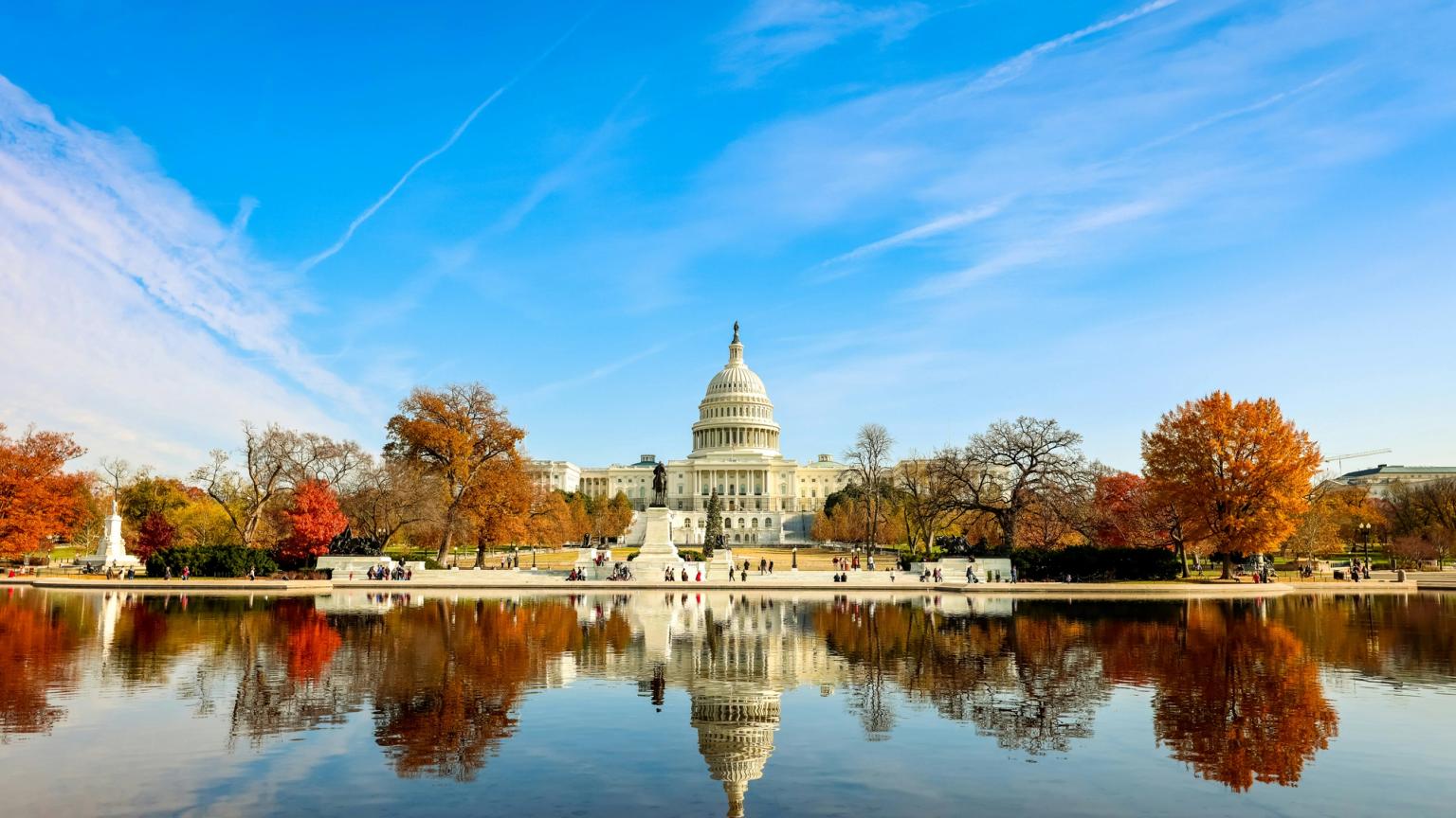 USA's Capitol-bygning spejler sig i et bassin med efterårstræer under en blå himmel.