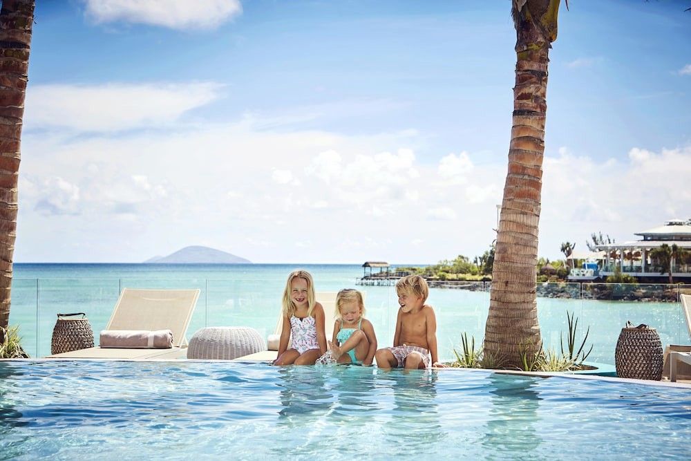 Tre små barn sitter vid kanten av en infinitypool med utsikt över ett tropiskt hav och en resort.