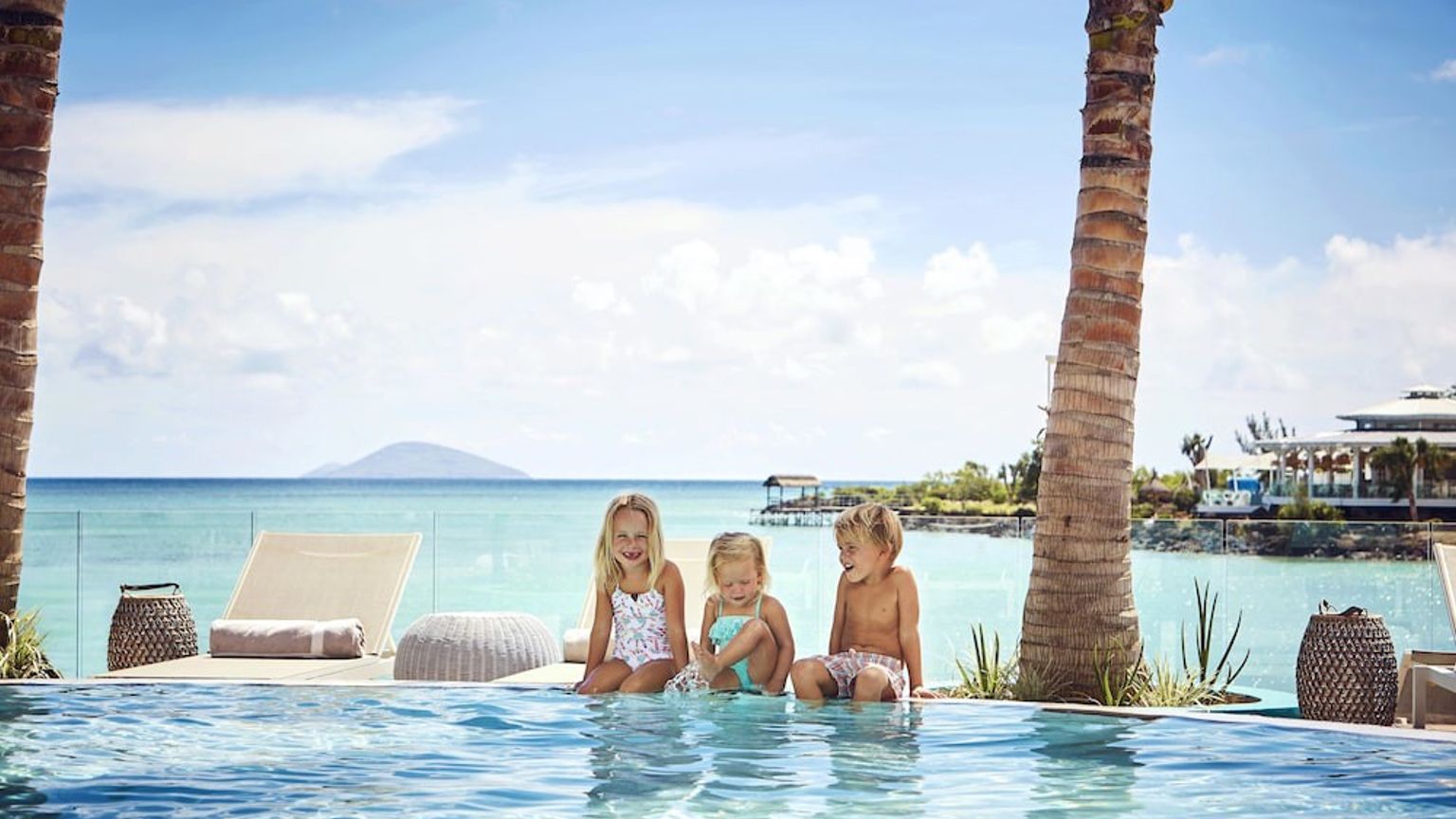 Tres niños pequeños sentados al borde de una piscina infinita con vistas a un océano tropical y un complejo turístico.