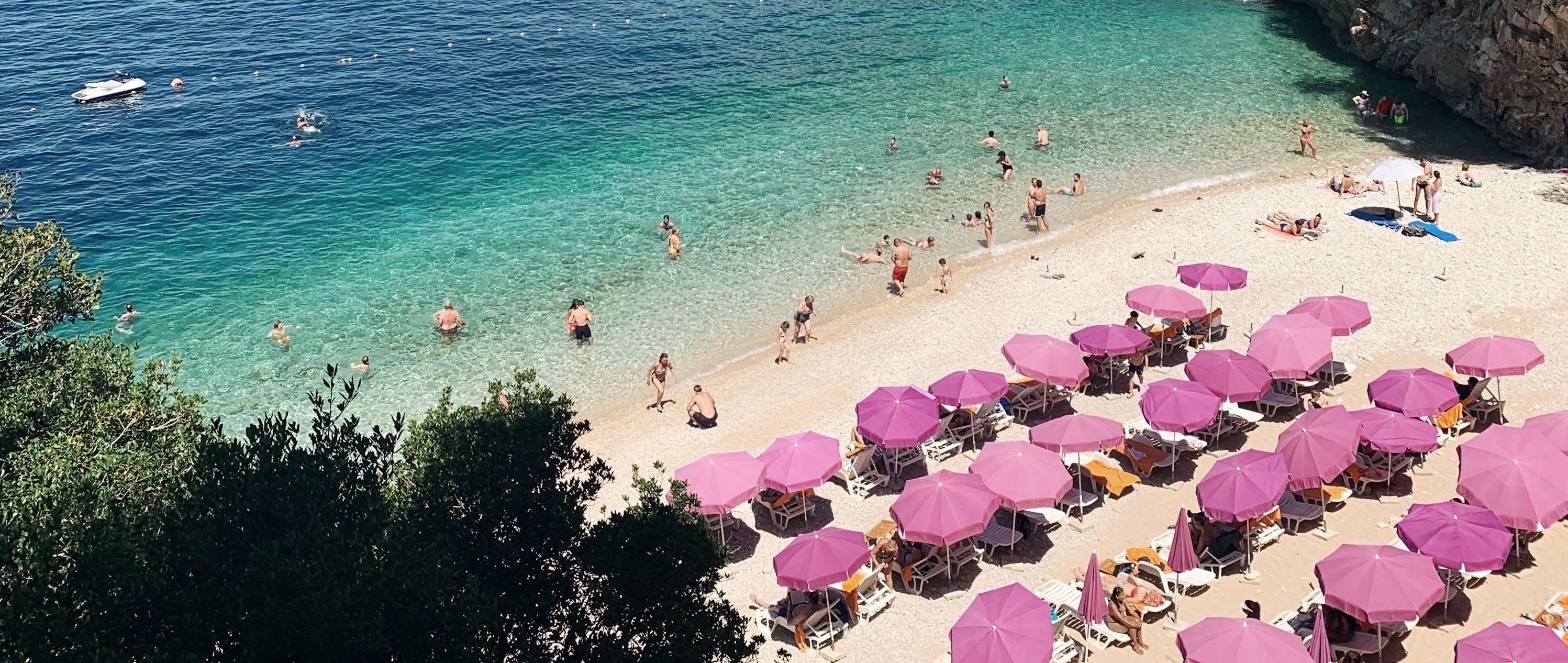 En fyldt strand med klart turkis vand, mange lyserøde parasoller og mennesker, der svømmer og solbader.