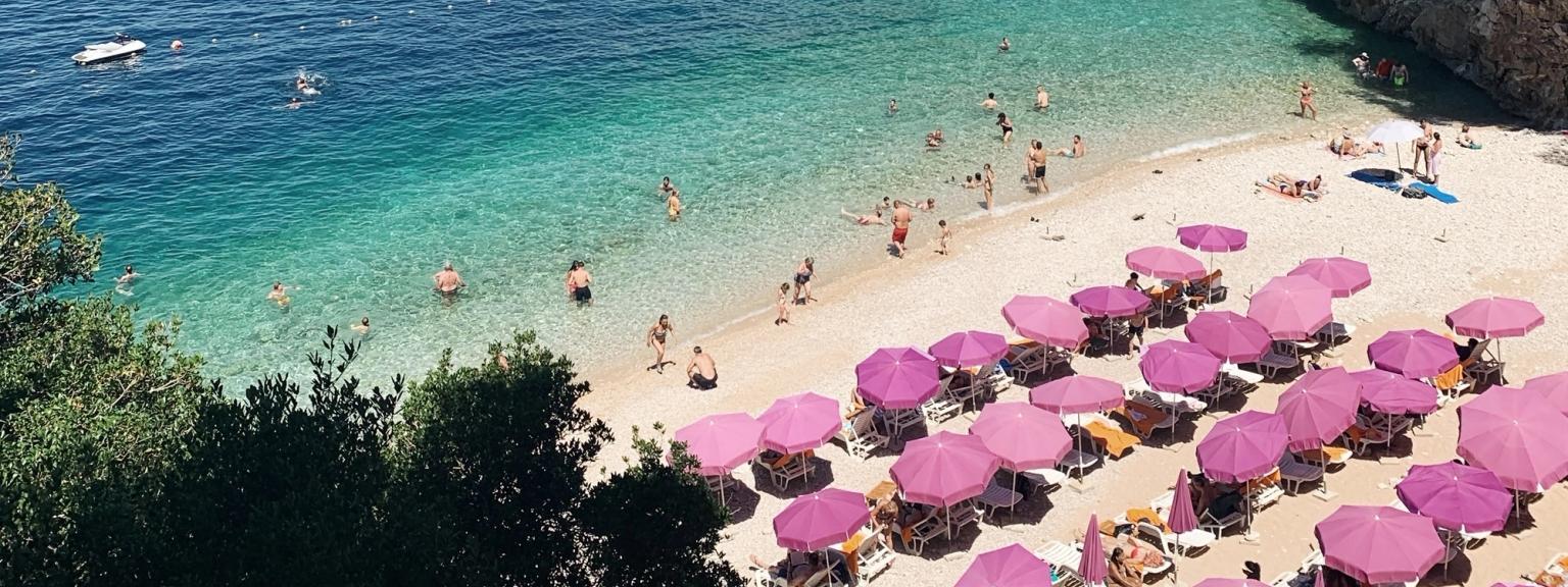 En fullpakket strand med klart turkis vann, mange rosa parasoller og folk som svømmer og soler seg.