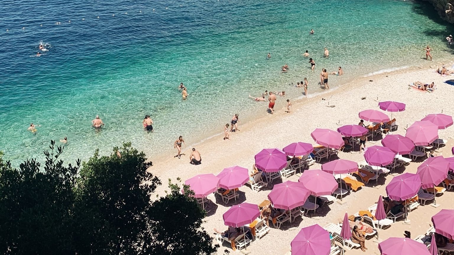 En fullpakket strand med klart turkis vann, mange rosa parasoller og folk som svømmer og soler seg.