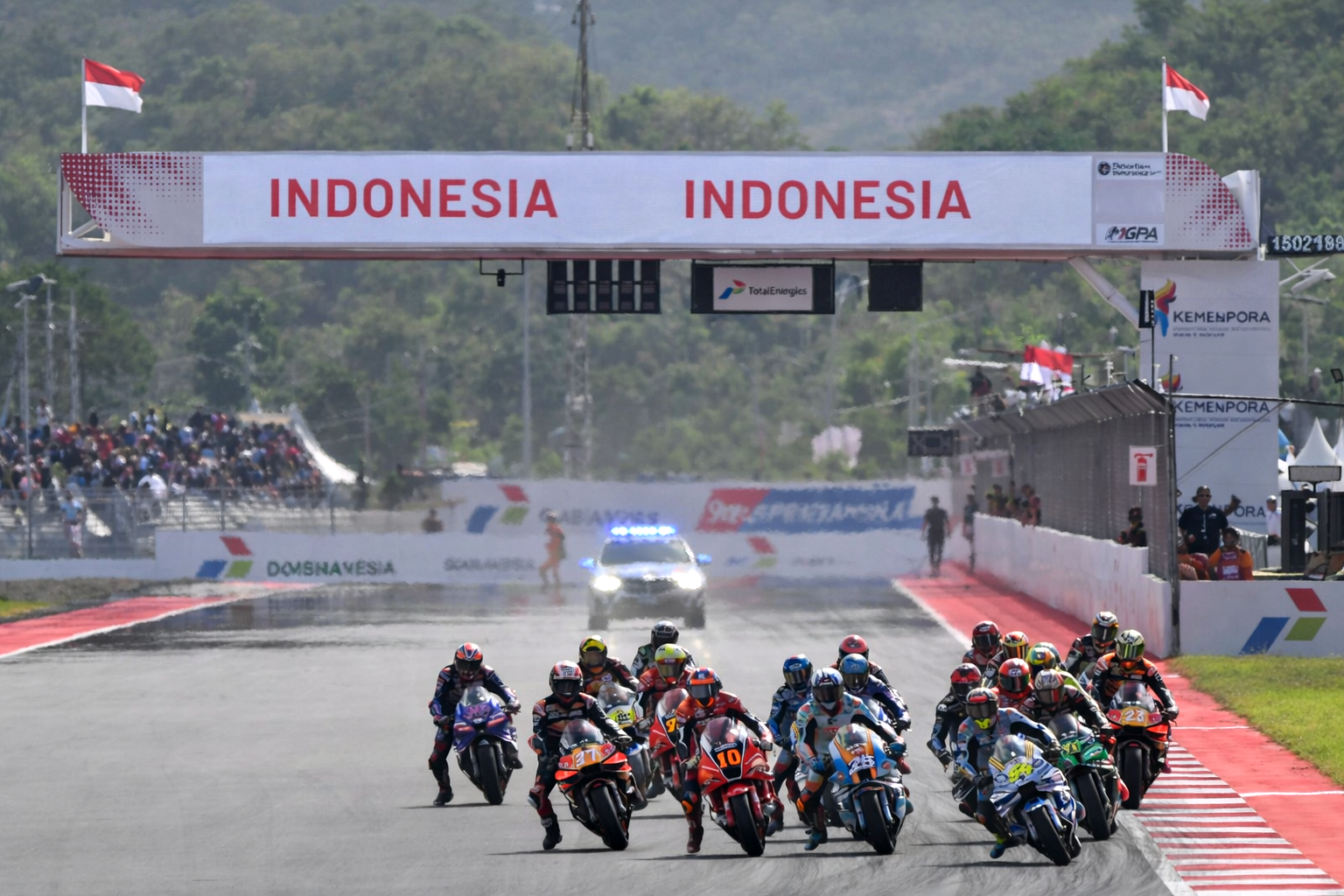 Motorcyclists race on a track under an "INDONESIA" banner, with a safety car visible.