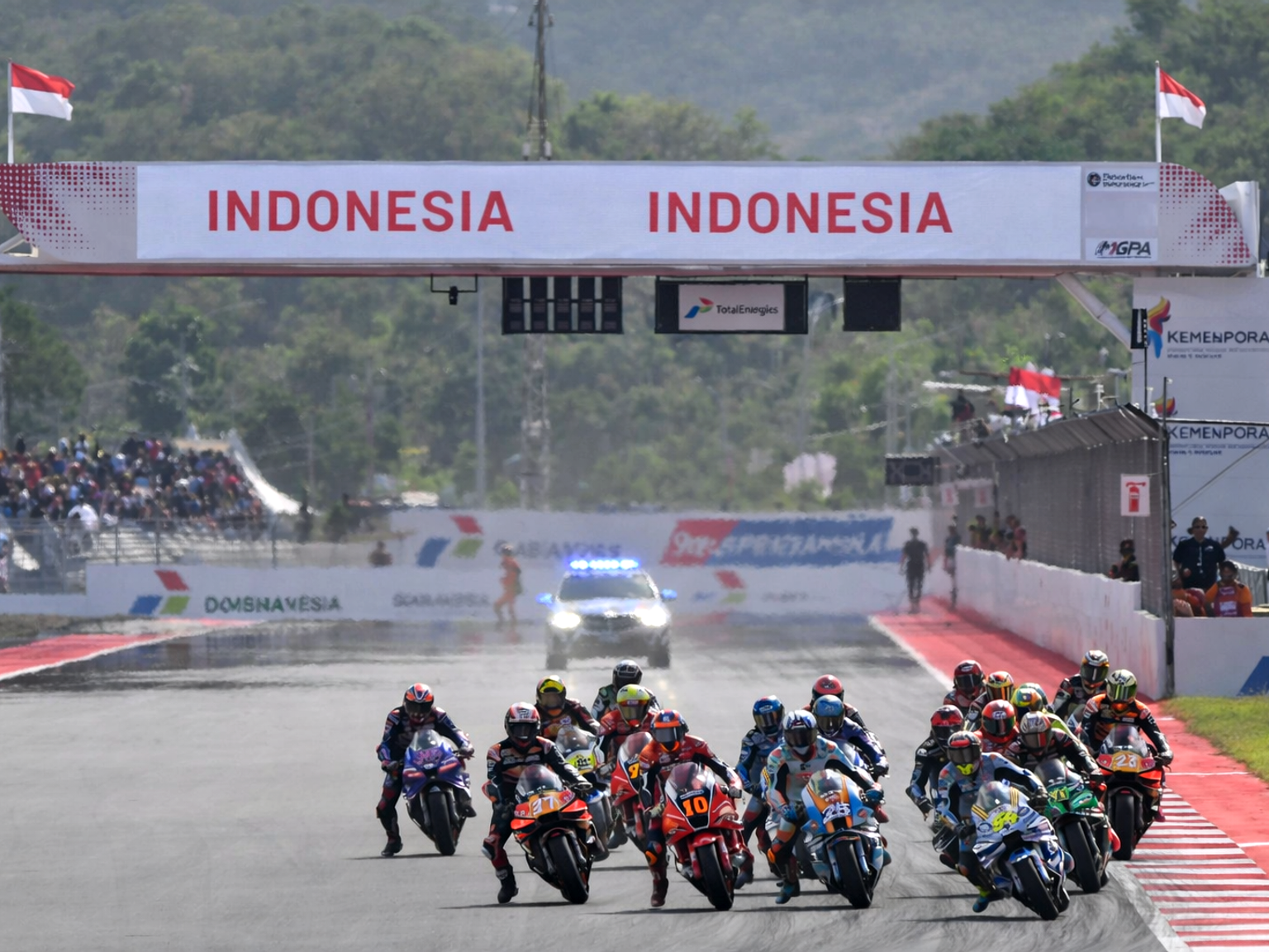 Motorcyclists race on a track under an "INDONESIA" banner, with a safety car visible.