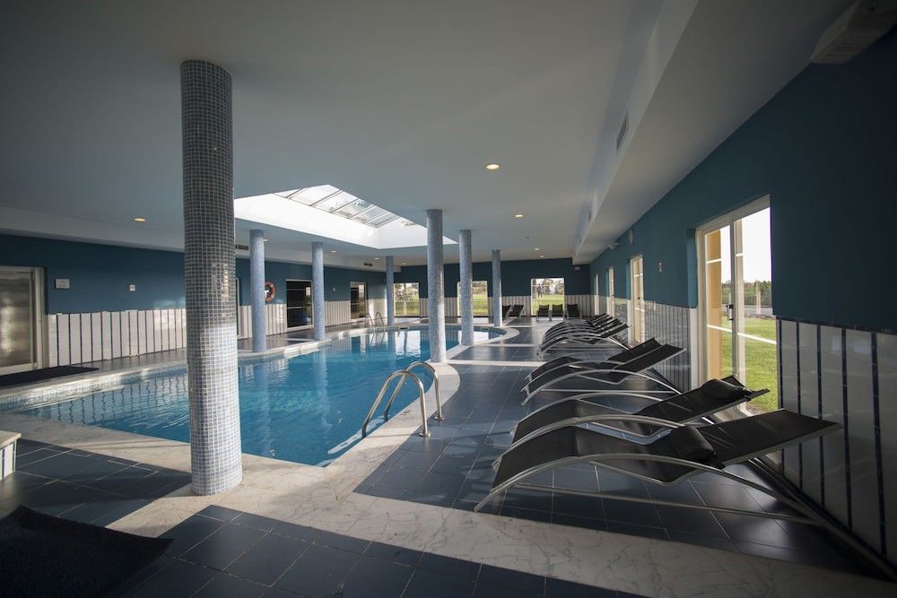Inomhuspool med blå väggar, kaklade pelare, ett takfönster och en rad solstolar.