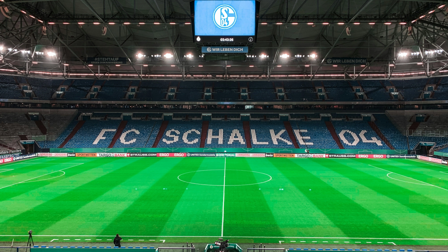 Ein leeres FC Schalke 04 Stadion mit einem grünen Spielfeld, blauen Sitzen, die den Teamnamen bilden, und einer großen Anzeigetafel mit ihrem Logo.