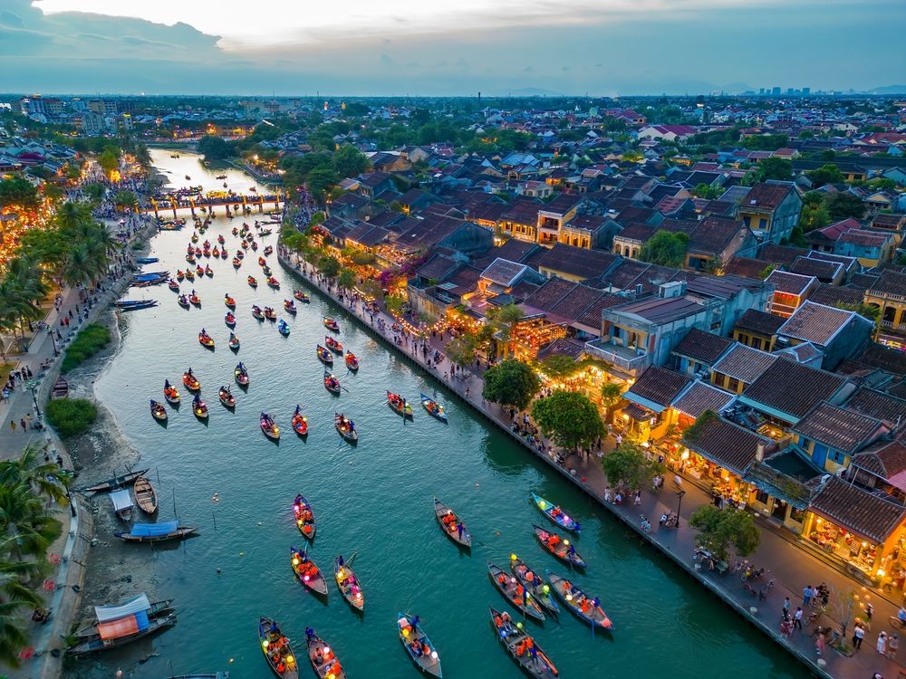 Aerial view of a vibrant riverside town at twilight, with numerous lantern boats on the water and illuminated buildings along the banks. Hoi An, Vietnam