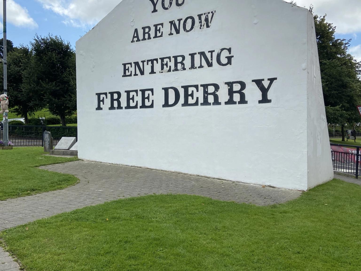 A white gable wall with black text 'YOU ARE NOW ENTERING FREE DERRY' standing in a grassy area with a paved path.