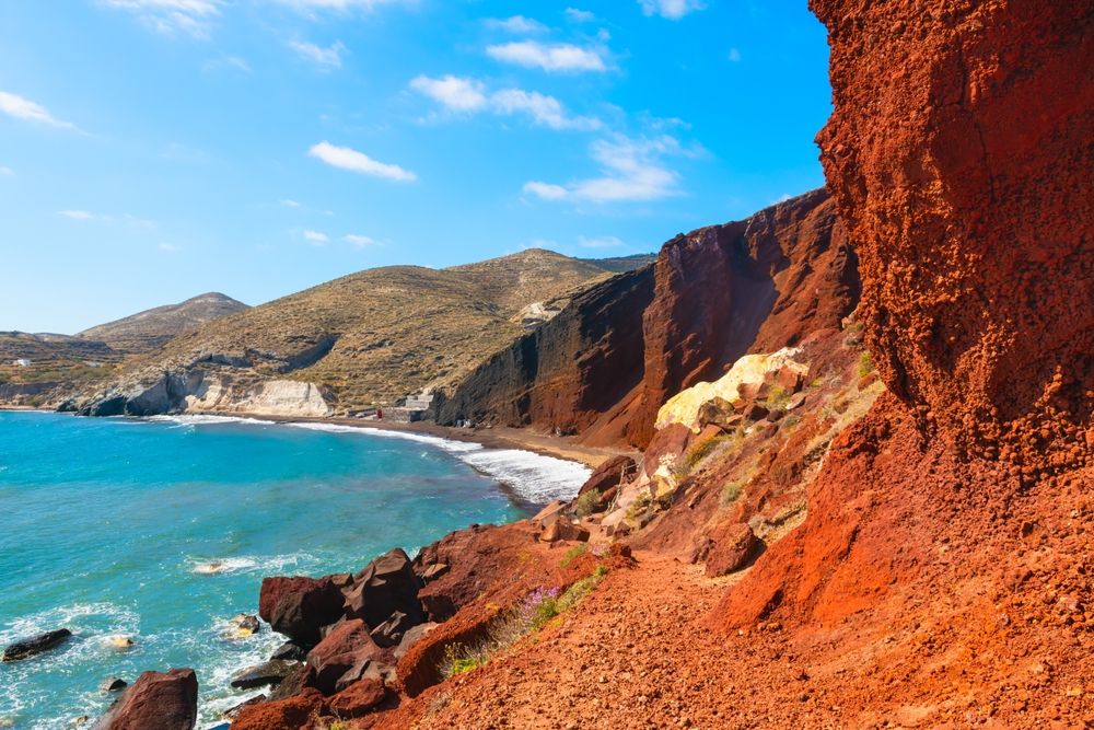 Red cliffs beside a turquoise bay with a dark sand beach and hills. Santorini, Greece