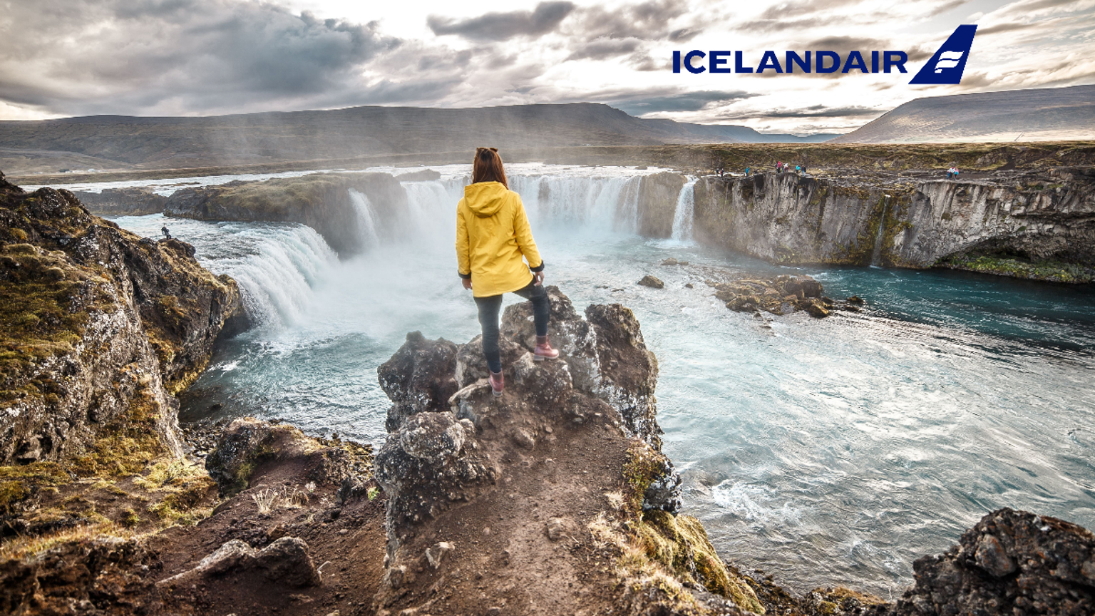 En kvinde i en gul jakke står på en klippe med udsigt over Goðafoss-vandfaldet, mens Icelandairs logo ses på himlen.