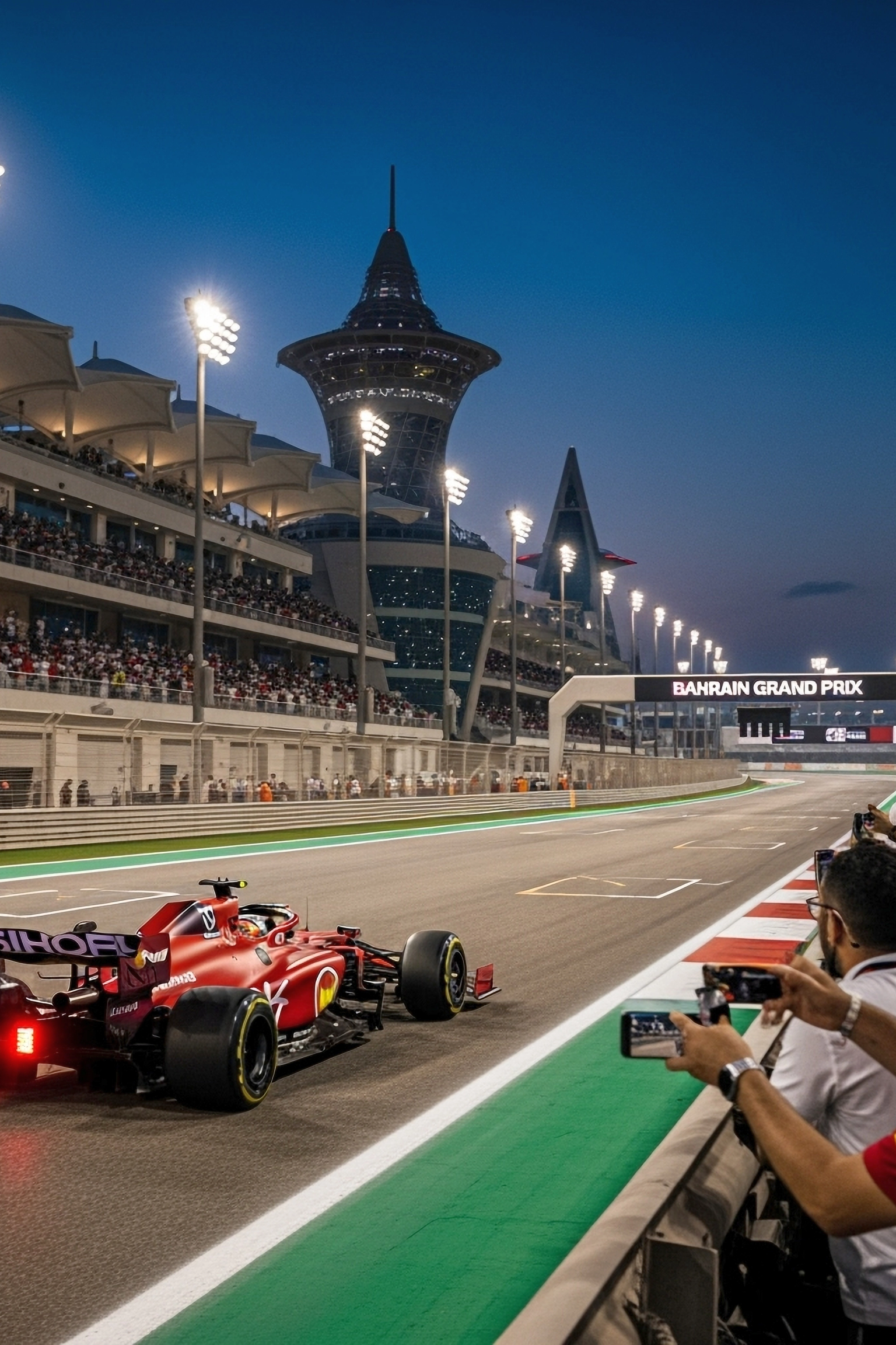 Punainen Formula 1 -auto, jonka takavalo hehkuu, ajaa valaistulla radalla yöllä Bahrainin Grand Prix -kilpailun aikana, katsomot täynnä yleisöä ja taustalla moderni torni.