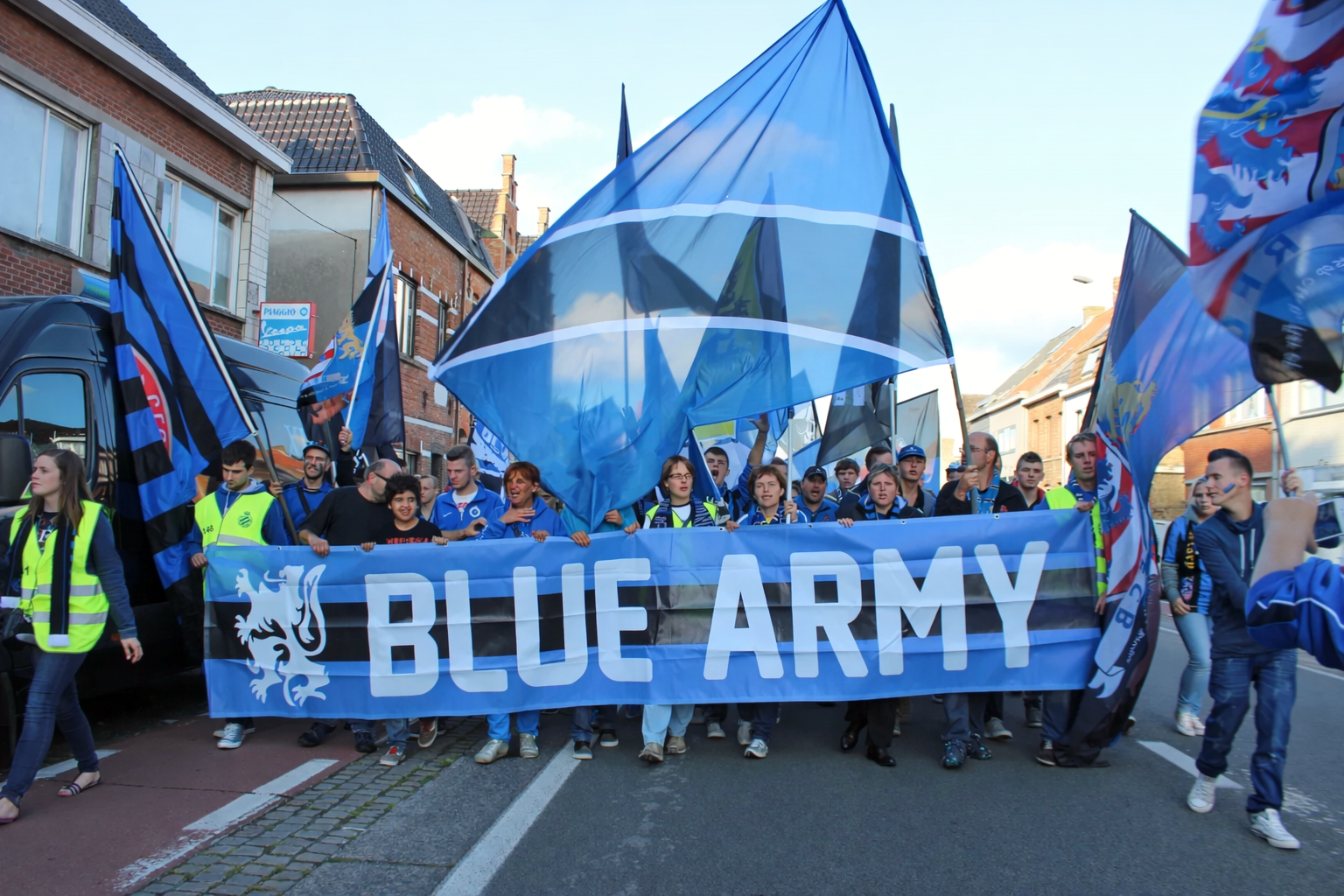 Ryhmä ihmisiä marssii kadulla kantaen lukuisia sinisiä lippuja ja banderollia, jossa lukee "BLUE ARMY" ja jossa on leijonaembleemi.