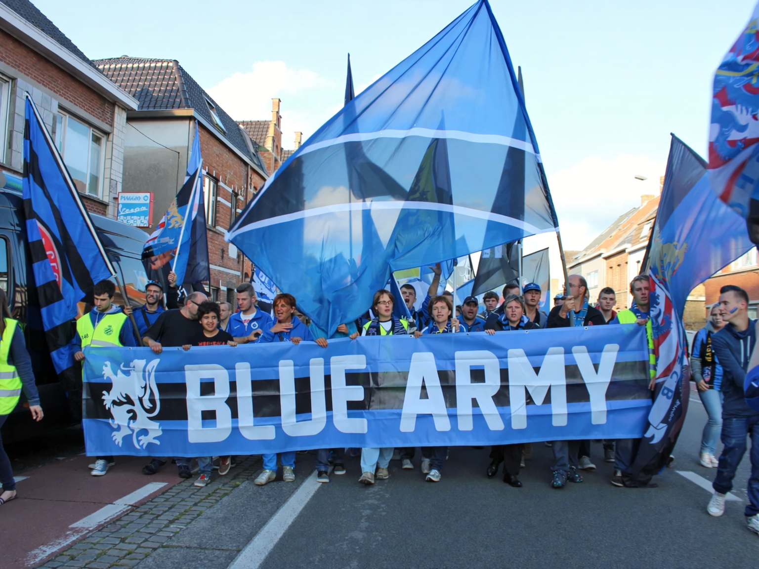Ryhmä ihmisiä marssii kadulla kantaen lukuisia sinisiä lippuja ja banderollia, jossa lukee "BLUE ARMY" ja jossa on leijonaembleemi.