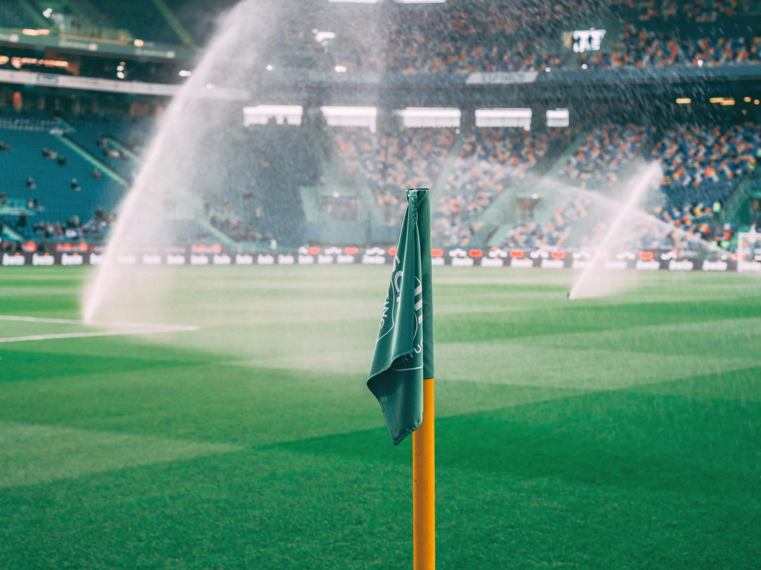 Green corner flag on a soccer field with sprinklers watering the grass and a stadium with spectators in the background.