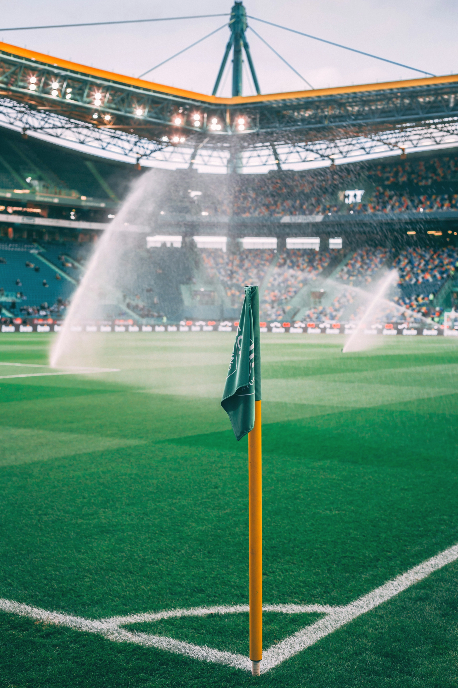 Green corner flag on a soccer field with sprinklers watering the grass and a stadium with spectators in the background.