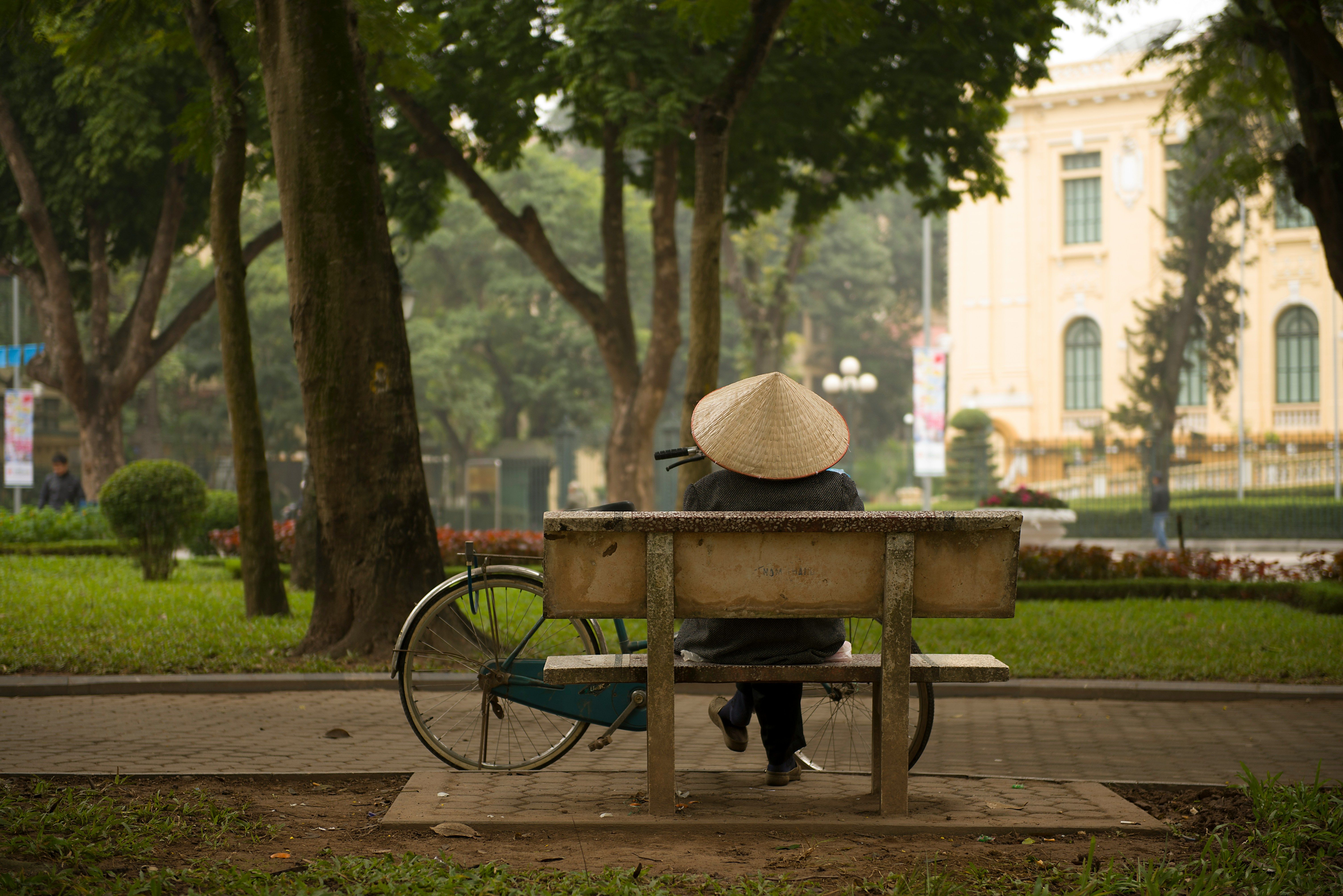 A person wearing a conical hat sits on a park bench next to a bicycle, with a large yellow building visible in the background. Hanoi, Vietnam