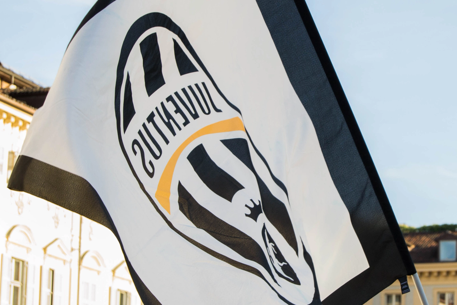 A Juventus flag with a mirrored logo waves in front of historic buildings under a clear sky.